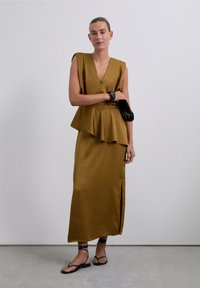 Senfgelbes Peplum-Oberteil mit tiefem V-Ausschnitt, kombiniert mit einem passenden langen Rock mit seitlichem Schlitz. Schwarze Clutch und römische Sandalen.