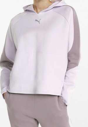 Woman wearing light purple and lavender Puma hoodie and matching sweatpants with hands in pockets, standing against white background.