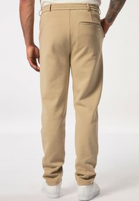 Beige Jogginghose mit elastischem Bund und einer Gesäßtasche, ausgestattet mit einer glatten Stofftextur und einem taillierten Bein-Design. Weiße Sneakers sind sichtbar.
