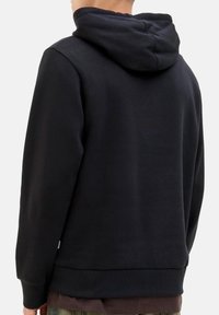 Person, der einen schlichten schwarzen Kapuzenpullover trägt, von hinten gezeigt, mit teilweise sichtbarem braunem Hemd und grüner Hose.