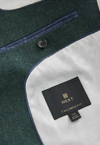 Jachetă din material verde închis, cu o căptușeală albă netedă, având un buton negru, o margine în nuanță de navy contrastantă și o etichetă ce indică "NEXT TAILORED FIT."
