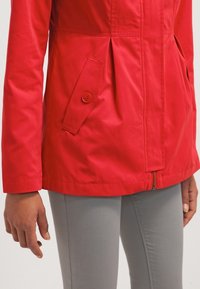 Veste rouge avec poches à rabat boutonnées et fermeture éclair à l'avant, portée avec un pantalon gris clair, montrant le bas du torse et les bras.