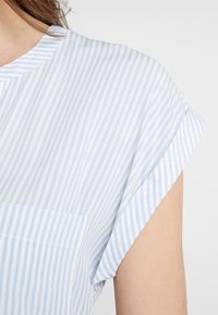 Blouse à rayures bleu clair et blanc avec un col rond, des manches courtes et une poche avant. Tissu doux avec une texture lisse.