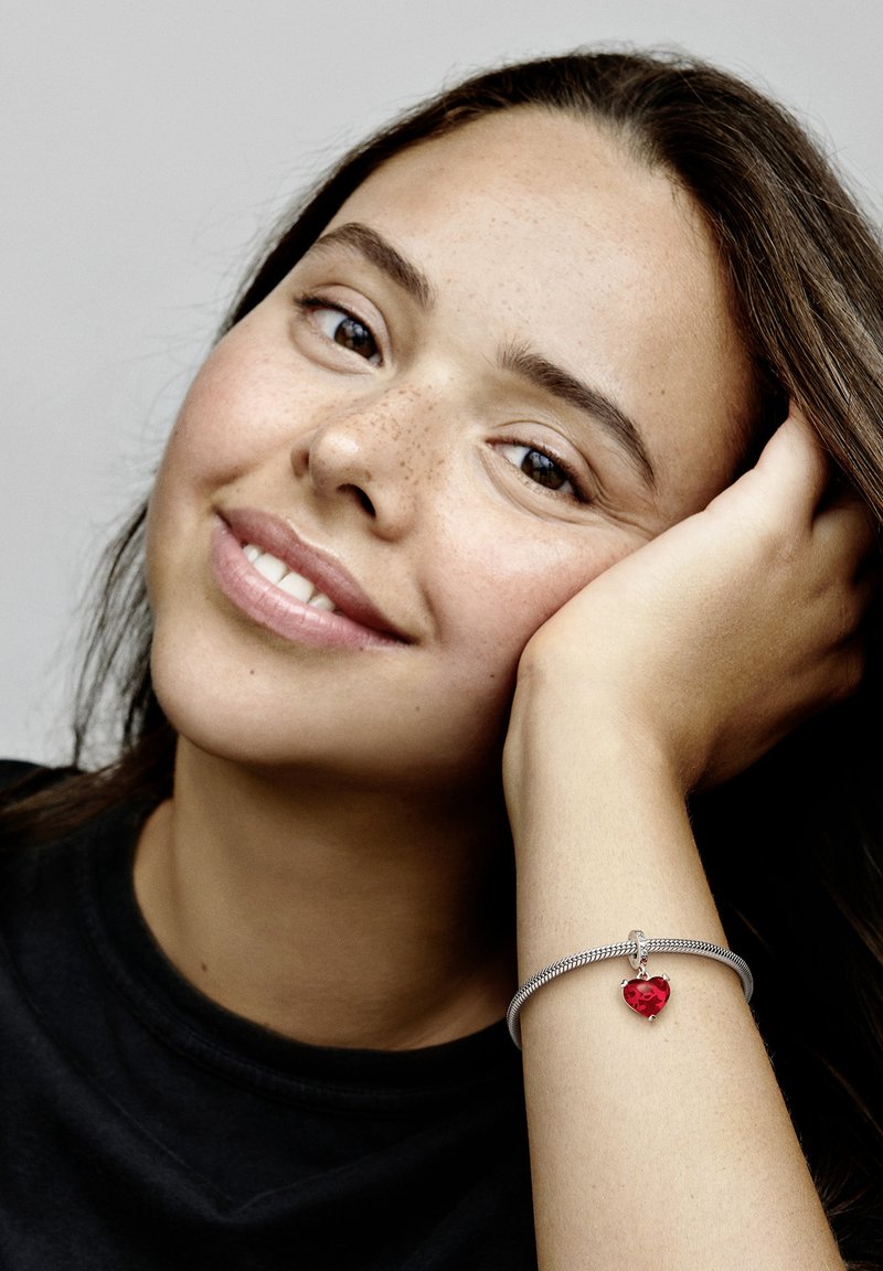 Bracciale d'argento con un ciondolo a forma di cuore rosso, caratterizzato da un design intrecciato e piccoli dettagli hardware, indossato su un polso appoggiato a un viso.