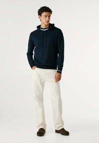 Mornarsko modra bombažna kapuca z vrvico, v kombinaciji s kremnimi hlačami. Opoznavna blagovna znamka na zapestju. Nosi se z rjavimi semiš čevlji.