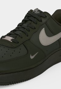 Σκούρο πράσινο αθλητικό παπούτσι Nike Air Force 1 με μαύρα κορδόνια και λευκά λογότυπα Nike Swoosh στο πλάι και στη γλώσσα.