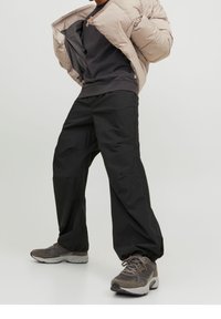 Vadderad beige jacka, svart sweatshirt och lösa svarta byxor; grå sneakers med snören; avslappnad oversized passform och texturerade material.