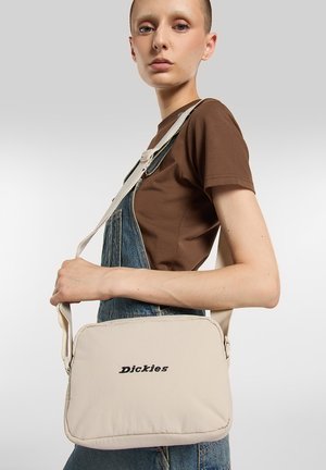 Beige polstret skuldertaske med broderet "Dickies"-logo, der har en aftagelig rem og en rektangulær form med afrundede kanter.