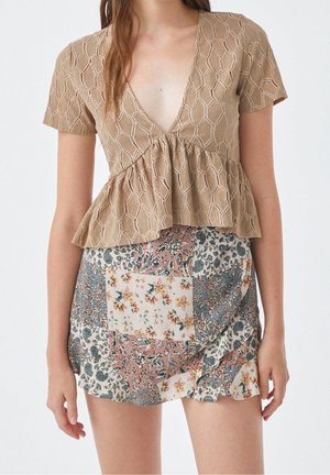 Femme portant un haut beige texturé à manches courtes avec un décolleté en V profond et une jupe patchwork florale multicolore, debout devant un fond uni.