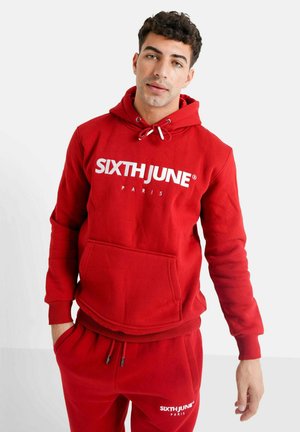 Sudadera con capucha roja, hecha de tela suave, con un bolsillo frontal y texto de logo en blanco. El diseño incluye cordones ajustables en la capucha.