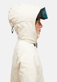 Person i beige hættejakke iført sorte og grønne skibriller, vist i profil mod en lys baggrund.