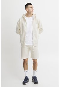 Lys beige hettegensere med snøring, parret med matchende shorts. Hvit t-skjorte under, svarte joggesko og hvite sokker. Enkel design.