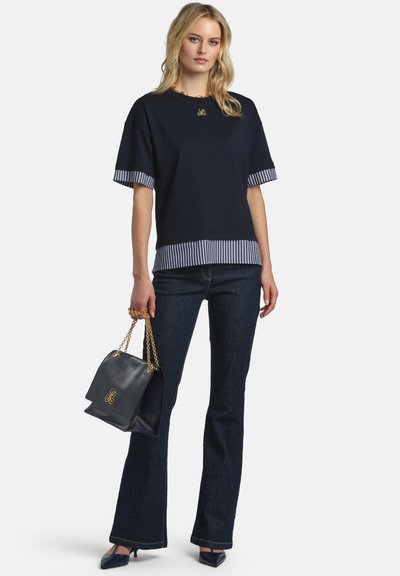 Woman wearing navy short-sleeve top with striped cuffs and hem, dark flared jeans, holding black handbag with gold chain straps.