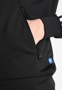 Svart sweatshirt med en ficka med dragkedja. Har ribbade muddar och nederkant. Liten blå Logotyp på sidan. Slät textur.