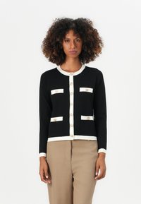 Svart cardigan med hvite detaljer, som har fire lommer og store gullknapper. Designet inkluderer rund hals og lange ermer.