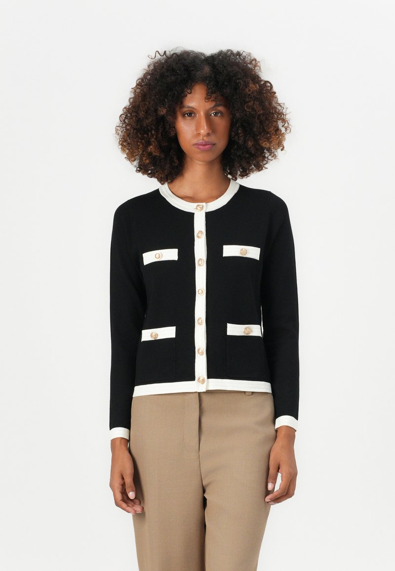 Svart cardigan med hvite detaljer, som har fire lommer og store gullknapper. Designet inkluderer rund hals og lange ermer.