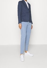 Blazer texturé marine, pantalons bleu clair et baskets blanches. Le blazer dispose d'un bouton unique et de deux poches avant.