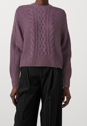 Kobieta ubrana w wrzosowy sweter z warkoczowym splotem, z prążkowanymi mankietami i dołem, w połączeniu z szerokimi czarnymi spodniami w prążki.