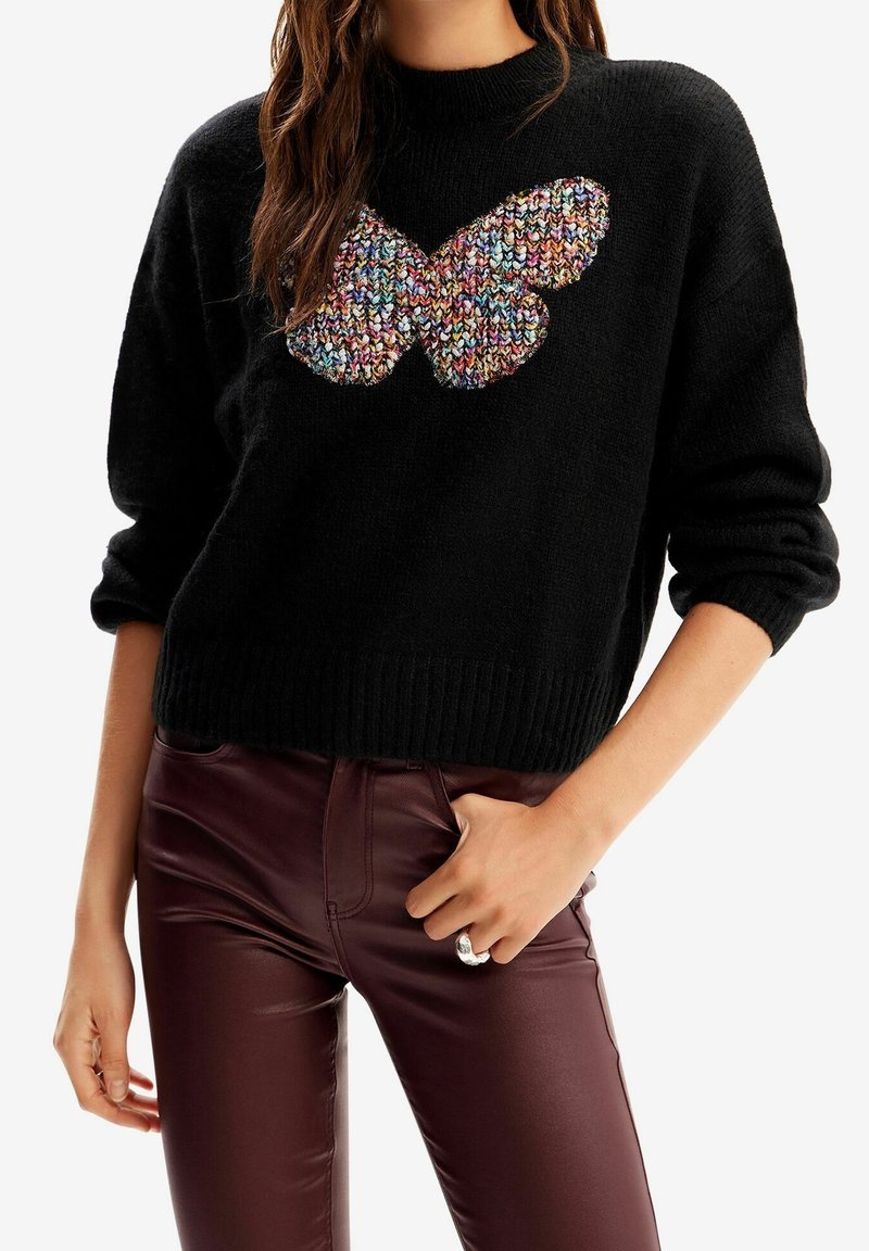 Femme portant un pull noir avec un motif papillon tricoté multicolore et un pantalon en cuir brun foncé, main reposant près de la poche.
