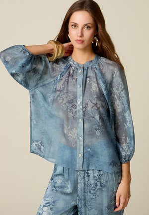 Vrouw die een doorschijnende blauwe bloemenblouse met knopen en bijpassende broek draagt, geaccentueerd met een grote gouden armband en oorbellen.