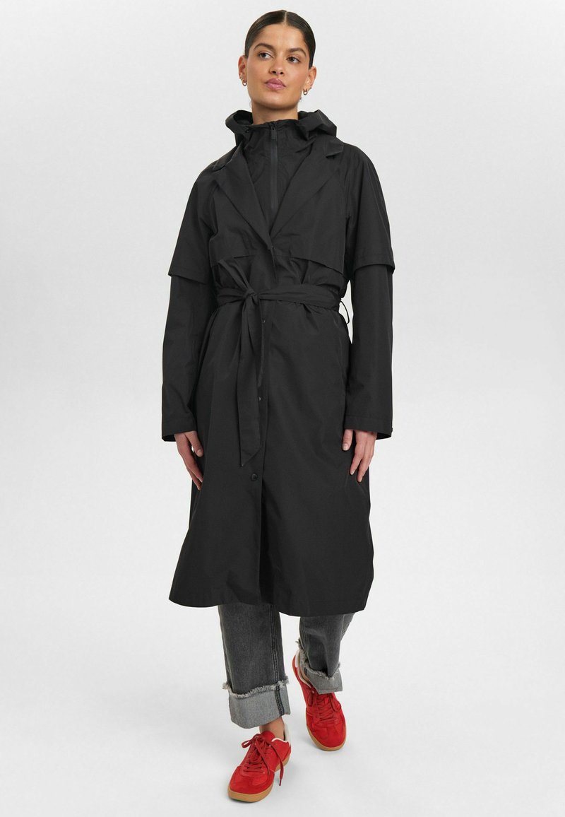 Svart vattentät trenchcoat med bälte i midjan, knäppning med knappar och huva; kombinerad med uppkavlade grå jeans och röda sneakers.