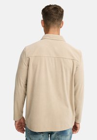 Mand iført en beige kordfløjljakke og blå denimjeans, stående med ryggen til kameraet mod en hvid baggrund.