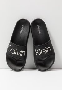 Paire de sandales slides noires Calvin Klein avec logo blanc sur les brides, semelles texturées, positionnées sur un fond blanc.