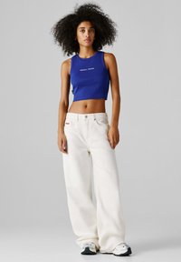 Nuori nainen kiharat hiukset, sininen lyhyt toppi ja väljävät valkoiset housut, seisoo yksiväristä harmaata taustaa vasten.