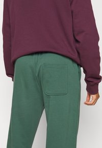 Gröna sweatpants med en bakficka, ihopparade med en djup lila sweatshirt. Materialet verkar mjukt; designen är avslappnad och bekväm.