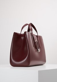 Bordeauxrode lederen handtas met een gestructureerd ontwerp, voorzien van twee handvatten en een zilverkleurig metalen detail aan de voorkant. Gladde textuur.