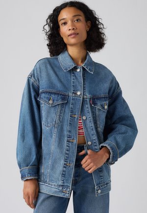 Donna con capelli ricci che indossa una giacca di jeans blu oversize sopra una maglia a righe rosse e bianche e jeans di jeans blu abbinati su uno sfondo semplice.