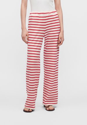 Personne portant un pantalon blanc ample avec des rayures rouges horizontales et des sandales noires à bouts ouverts, debout devant un fond blanc uni.
