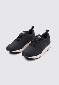 Zapatillas deportivas negras con parte superior sintética suave, punta redonda, diseño de cordones y una suela media blanca que presenta un logo en el costado.