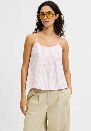 Vrouw draagt een wit mouwloos topje met fijne strepen, beige geplooide broek en grote geel getinte zonnebril, staat met één hand in de zak.