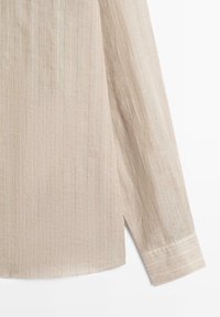 Beżowa tkanina koszuli z długim rękawem z cienkimi pionowymi białymi paskami i wykończeniem mankietu z przeszyciami na jednolitym tle.