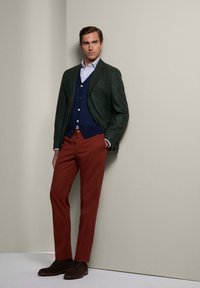 Man die leunt tegen een beige muur, gekleed in een donkergroene blazer, een marineblauwe cardigan, een geruit overhemd, roestkleurige broek en donkerbruine schoenen.