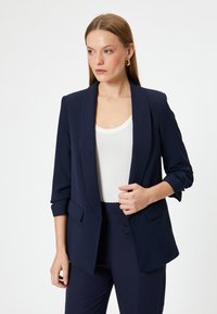 Blazer navy con solapa de chal, cierre de un solo botón y bolsillos frontales. La tela tiene un acabado suave y un diseño estructurado.