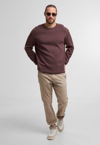 Bordeauxfarbener Pullover, beige Hose und weiße Sneakers mit strukturierten Details. Die Person trägt runde Sonnenbrillen und steht selbstbewusst.