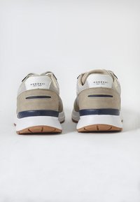 Beige és navy színű sportcipők, texturált felsőrésszel, hálós panelekkel és gumitalppal. A saroknál a márka logója, valamint egyedi dizájn elemek találhatók.