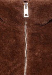 Veste en fausse fourrure marron avec une texture lisse, dotée d'une fermeture éclair argentée au centre et d'un col arrondi.