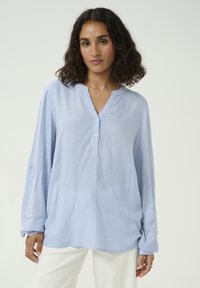 Lichtblauw langarmshirt met een V-hals en drie knopen. Heeft opgerolde mouwen en een zachte, textuur stof. Witte achtergrond.