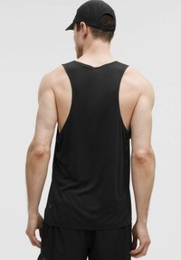 Uomo che indossa una canotta atletica senza maniche nera e un cappellino nero, in piedi con la schiena rivolta verso la camera su uno sfondo chiaro uniforme.
