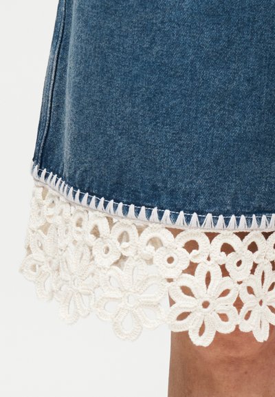 Παντελονάκι από denim με λευκή φλοράλ δαντέλα και διακοσμητικό ράψιμο, εμφανιζόμενο στο πόδι ενός ατόμου σε απλό φόντο.