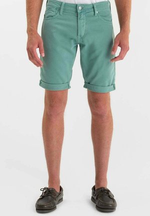 Uomo che indossa pantaloncini turchese risvoltati e scarpe da barca in pelle marrone scuro, in piedi su sfondo bianco.