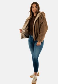 Chaqueta de piel de imitación marrón con un revestimiento interior brillante, que cuenta con una capucha y cremallera frontal. Lleva puesta sobre un suéter marrón oscuro y jeans azules.