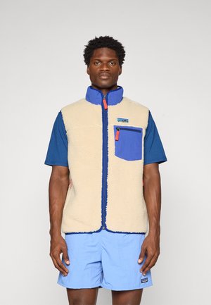 Fleece vest in beige with blue accents, zip closure, high collar, front pocket, worn over a blue t-shirt and light blue shorts.