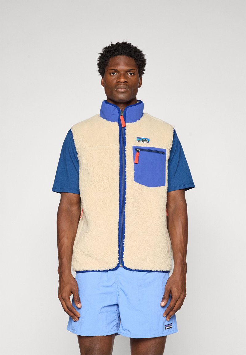 Fleece vest in beige with blue accents, zip closure, high collar, front pocket, worn over a blue t-shirt and light blue shorts.