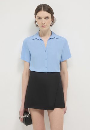 Chemise bleu clair à manches courtes avec boutons, associée à une mini-jupe portefeuille noire. Le mannequin tient un petit sac noir avec une bandoulière chaîne argentée.