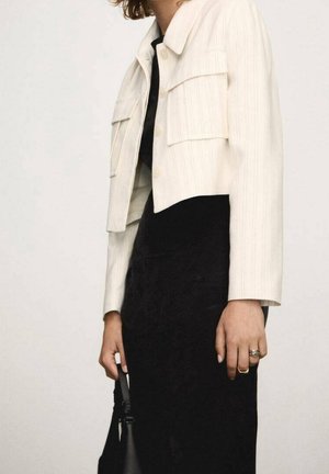 Person wearing a cropped white jacket over a black dress, holding a black handbag, with rings on fingers visible.