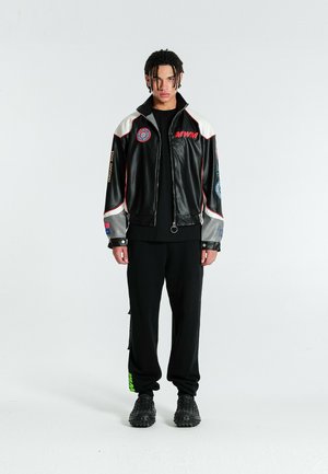 Joven con el pelo trenzado lleva una chaqueta con cremallera en blanco y negro con parches, pantalones negros y zapatos negros texturizados, de pie contra un fondo blanco liso.